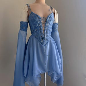 Puede incluir: Un vestido azul claro con un corpi&ntilde;o estilo cors&eacute; y una falda fluida. El corpi&ntilde;o est&aacute; adornado con intrincados abalorios y un frente con cordones. El vestido presenta mangas largas acampanadas y delicadas tiras finas.