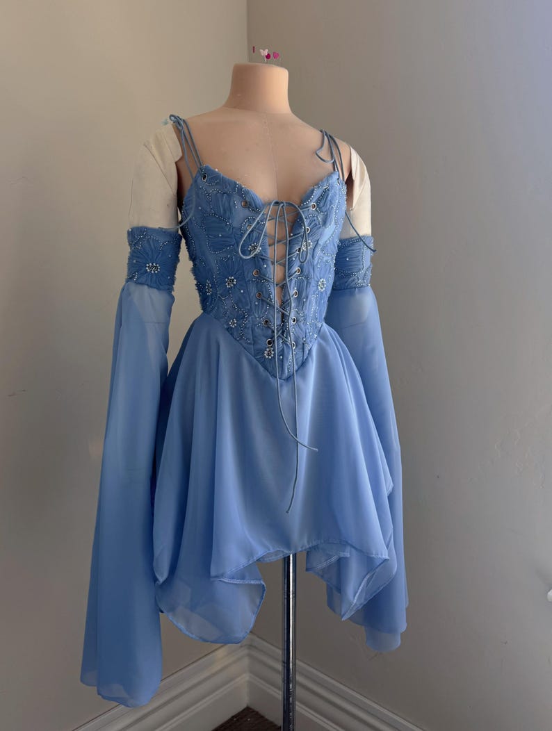 Puede incluir: Un vestido azul claro con un corpi&ntilde;o estilo cors&eacute; y una falda fluida. El corpi&ntilde;o est&aacute; adornado con intrincados abalorios y un frente con cordones. El vestido presenta mangas largas acampanadas y delicadas tiras finas.