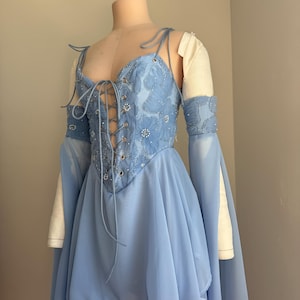 Puede incluir: Un vestido azul claro con un corpi&ntilde;o estilo cors&eacute; y una falda fluida. El corpi&ntilde;o est&aacute; adornado con bordados florales y adornos brillantes. El vestido presenta mangas largas y fluidas y se exhibe en un maniqu&iacute;.