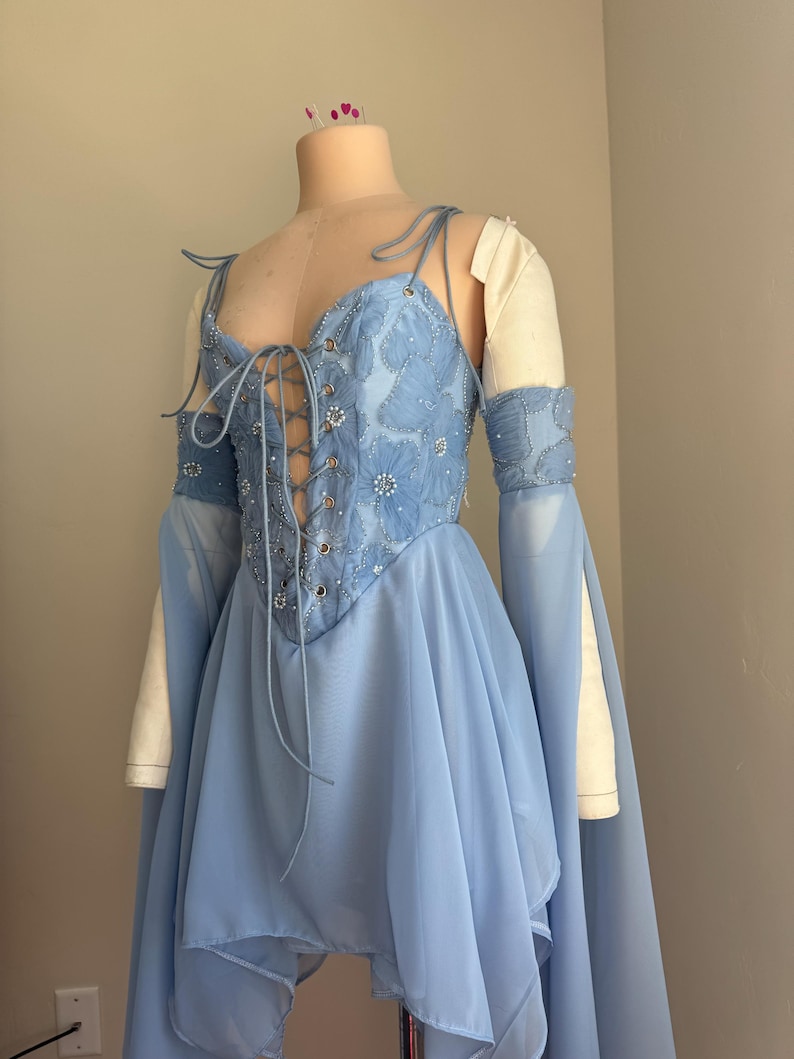 Puede incluir: Un vestido azul claro con un corpi&ntilde;o estilo cors&eacute; y una falda fluida. El corpi&ntilde;o est&aacute; adornado con bordados florales y adornos brillantes. El vestido presenta mangas largas y fluidas y se exhibe en un maniqu&iacute;.