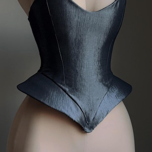 Puede incluir: Un cors&eacute; gris oscuro con un dise&ntilde;o estructurado y angular. El corpi&ntilde;o tiene un escote en forma de V y una cintura estilo peplum. La tela tiene un brillo sutil, y el cors&eacute; se muestra en un maniqu&iacute;.