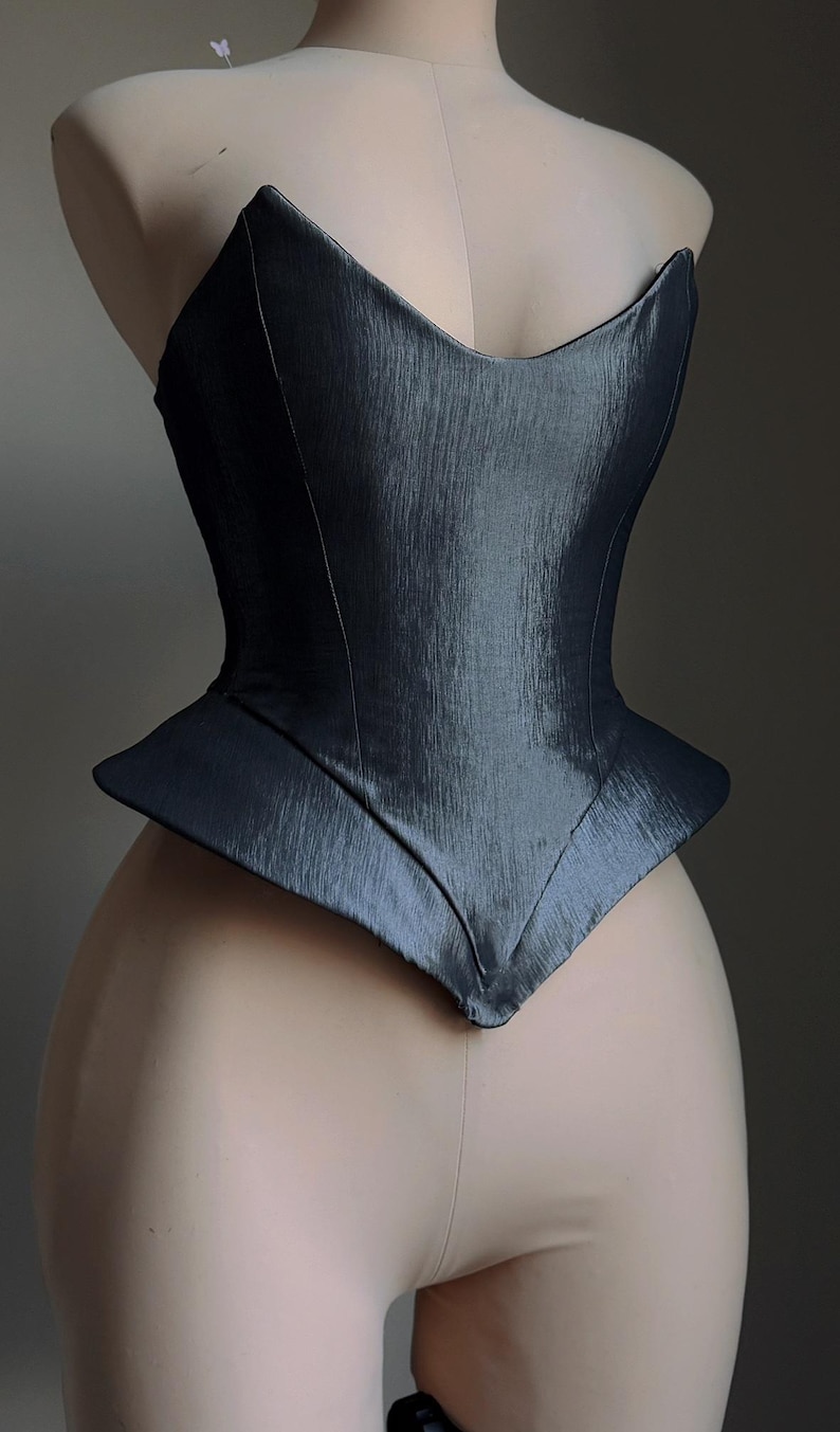 Puede incluir: Un cors&eacute; gris oscuro con un dise&ntilde;o estructurado y angular. El corpi&ntilde;o tiene un escote en forma de V y una cintura estilo peplum. La tela tiene un brillo sutil, y el cors&eacute; se muestra en un maniqu&iacute;.
