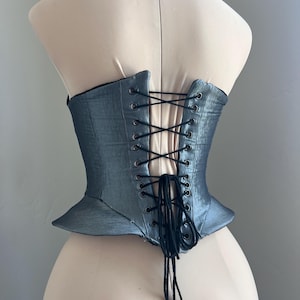 Puede incluir: Un cors&eacute; azul acero con una silueta estructurada. La parte trasera presenta un cierre de cordones con cord&oacute;n negro y ojales met&aacute;licos. El cors&eacute; se exhibe en un maniqu&iacute;.