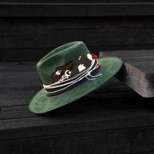 May include: A green felt fedora hat with a white and black design on the crown. The hat has a black leather band with a white cord and a red tassel.