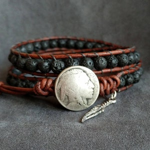 Pu&ograve; includere: Un braccialetto avvolgente in pelle marrone con perline di lava nera e un ciondolo in argento a forma di nichel di bufalo. Un ciondolo in argento a forma di piuma pende dal braccialetto.