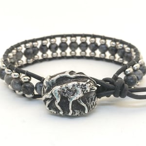 May include: A wrap bracelet featuring gray and silver beads, a black leather band, and a silver wolf charm. The bracelet is designed with a double wrap and a knotted closure. The wolf charm is detailed with a textured finish.