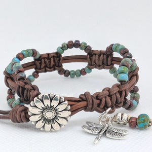 May include: A brown leather bracelet with turquoise and maroon beads. The bracelet features a silver daisy clasp and a dragonfly charm. The macrame design is accented with small beads, creating a bohemian style.