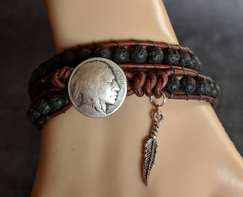 Pu&ograve; includere: Un braccialetto in pelle marrone con perline di lava nera e un ciondolo a forma di moneta da nickel con la testa di un indiano in argento. Un ciondolo a forma di piuma d'argento &egrave; appeso al braccialetto.