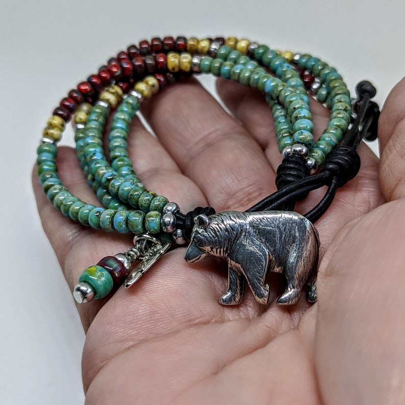 May include: A multi-strand beaded bracelet with a silver bear charm. The bracelet is made with turquoise, yellow, and red beads. The bear charm is attached to a black leather cord.
