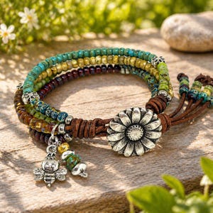 May include: A multi-strand beaded bracelet with a brown leather wrap and a silver-tone sunflower clasp. The bracelet features turquoise, yellow, and brown beads, with a bee charm and other decorative beads.