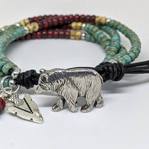 May include: A multi-strand bracelet with turquoise, red, and beige beads. The bracelet has a silver bear charm and a silver arrowhead charm.