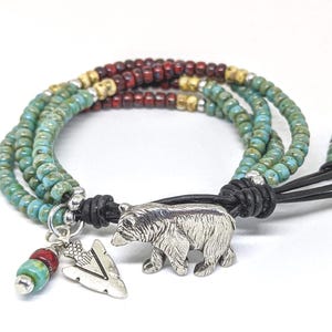 May include: Multi-strand bracelet with turquoise, red, and gold beads. The bracelet has a leather closure and a silver bear charm with an arrowhead charm.