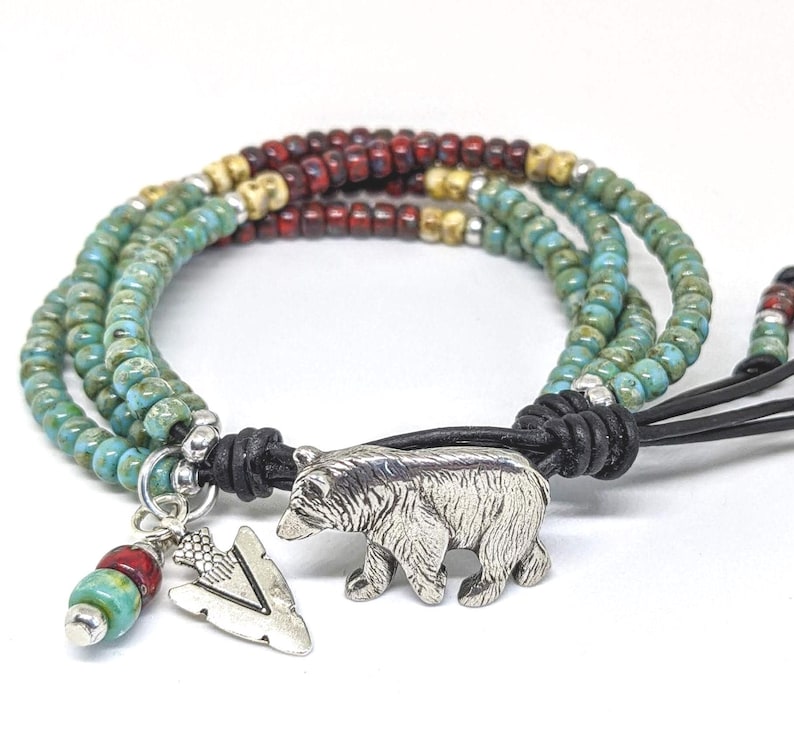 May include: Multi-strand bracelet with turquoise, red, and gold beads. The bracelet has a leather closure and a silver bear charm with an arrowhead charm.