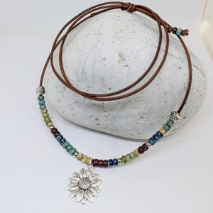 May include: A necklace with a brown leather cord and a silver sunflower pendant. The necklace features a strand of colorful beads in shades of blue, green, yellow, and red. The necklace is displayed on a white stone.