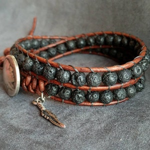 Pu&ograve; includere: Un braccialetto in pelle marrone con perline di lava nera e un ciondolo a forma di piuma d'argento.