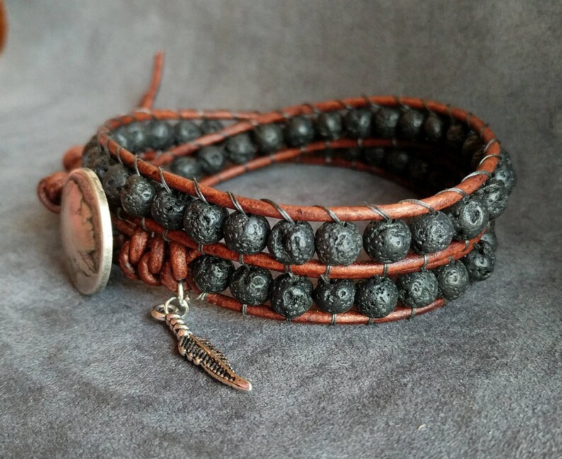 Pu&ograve; includere: Un braccialetto in pelle marrone con perline di lava nera e un ciondolo a forma di piuma d'argento.