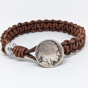 May include: Brown leather bracelet with a braided design and a silver coin featuring a Native American profile. The bracelet has a decorative silver bead and a clasp closure. A rustic accessory.
