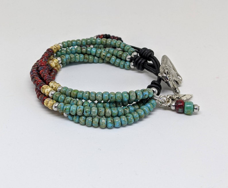 May include: Multi-strand bracelet with turquoise, cream, and red beads. The bracelet has a leather closure with a silver charm and a feather dangle.