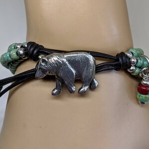May include: A leather bracelet with a silver bear charm and turquoise and red beads.