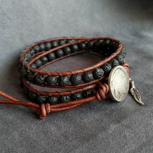 Pu&ograve; includere: Un braccialetto in pelle marrone con perline di lava nera e un ciondolo a forma di moneta d'argento con un accento di piuma.