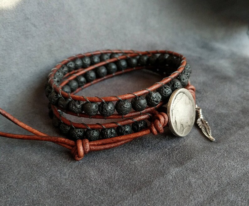 Pu&ograve; includere: Un braccialetto in pelle marrone con perline di lava nera e un ciondolo a forma di moneta d'argento con un accento di piuma.