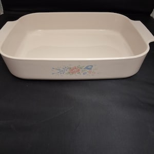 May include: Off-white rectangular baking dish with handles. The dish has a floral design in the lower center with pink, blue, and green accents. The dish is empty and sits on a dark surface.