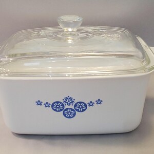 May include: White rectangular casserole dish with a clear glass lid and handle. The dish features a blue floral pattern on the front. The lid has a clear knob handle. A classic kitchenware item.