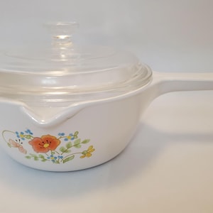 May include: White saucepan with a clear glass lid and a long handle. The side of the pan features a floral design with orange, blue, and yellow flowers. The pan has a pouring spout.