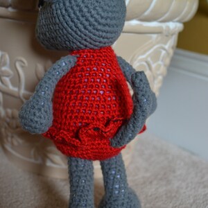 May include: A gray crocheted mouse wearing a red dress with a ruffled hem. The mouse is standing on a light brown surface.
