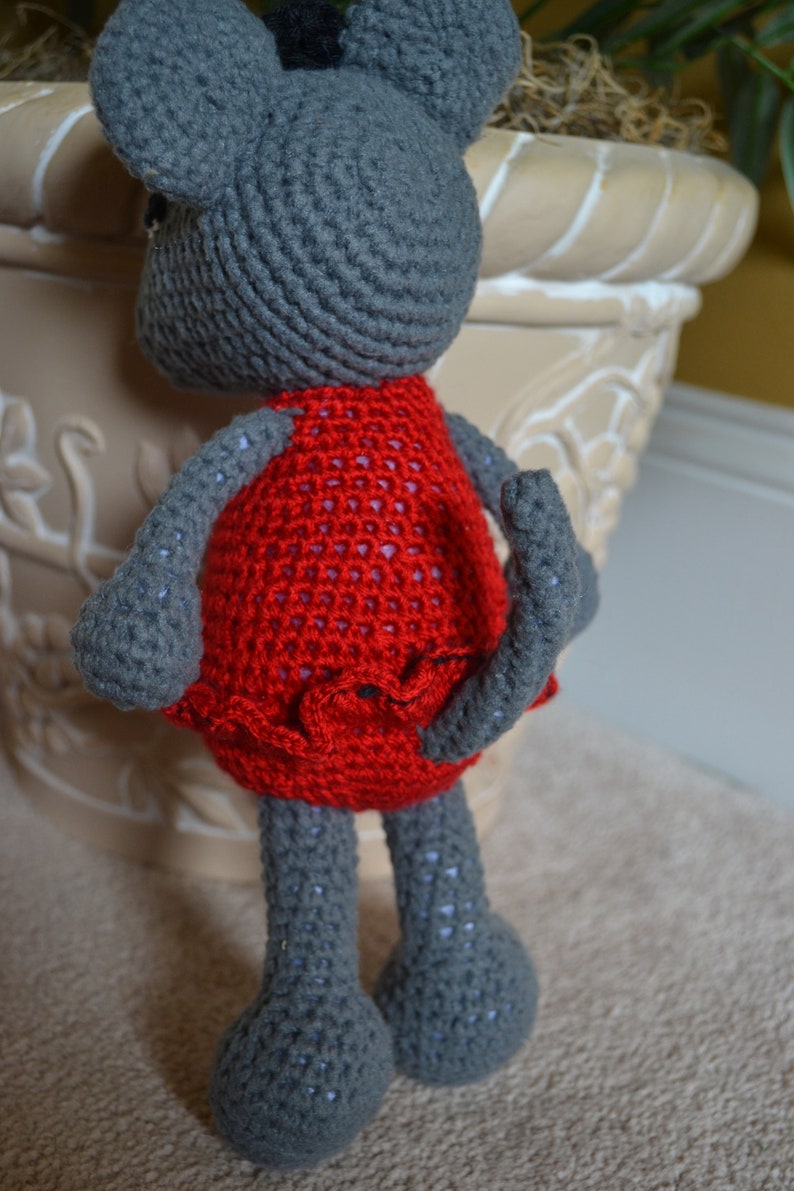 May include: A gray crocheted mouse wearing a red dress with a ruffled hem. The mouse is standing on a light brown surface.