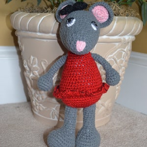 May include: A gray crocheted mouse wearing a red dress with a black beret. The mouse is standing on a light brown surface.
