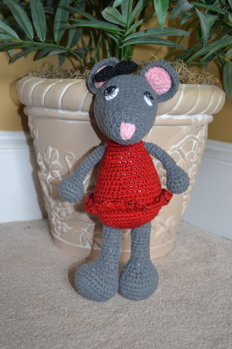 May include: A gray crocheted mouse wearing a red dress with a black beret. The mouse is standing on a light brown surface.