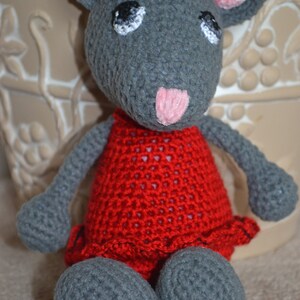 May include: A gray crocheted mouse wearing a red dress with a black bow. The mouse has pink ears and a pink nose.