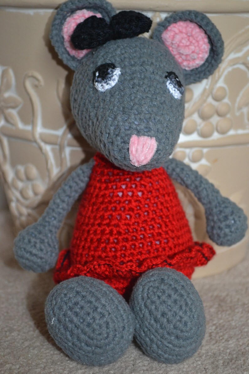 May include: A gray crocheted mouse wearing a red dress with a black bow. The mouse has pink ears and a pink nose.