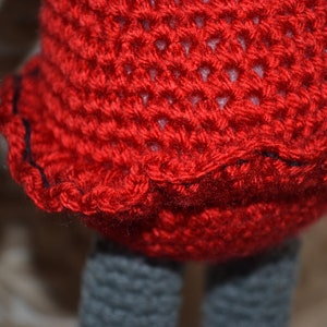 May include: A crocheted doll with a red dress and gray legs. The dress has a black trim around the bottom.