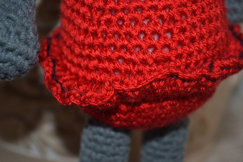 May include: A crocheted doll with a red dress and gray legs. The dress has a black trim around the bottom.