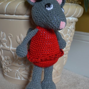 May include: A gray crocheted mouse wearing a red dress with a black bow. The mouse is standing next to a white planter with a floral design.