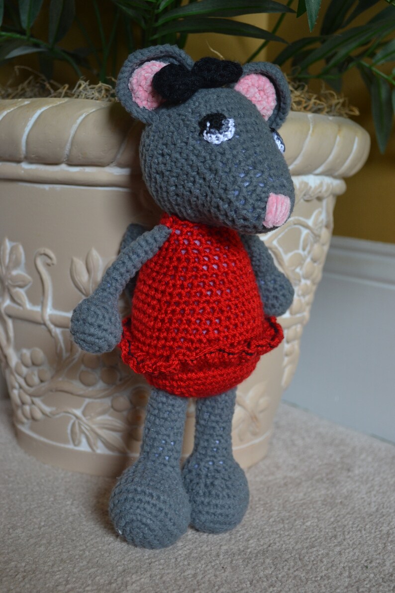 May include: A gray crocheted mouse wearing a red dress with a black bow. The mouse is standing next to a white planter with a floral design.