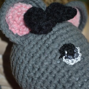 May include: A gray crocheted mouse toy with black yarn ears and a pink nose. The mouse has a black eye with a white outline and is wearing a red crocheted scarf.