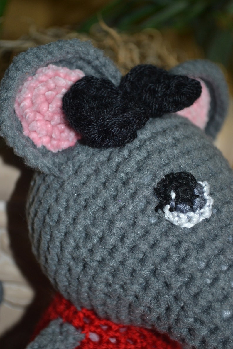 May include: A gray crocheted mouse toy with black yarn ears and a pink nose. The mouse has a black eye with a white outline and is wearing a red crocheted scarf.