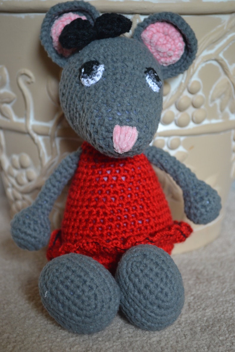 May include: A gray crocheted mouse wearing a red dress with a black bow on its head. The mouse is sitting on a light brown surface.