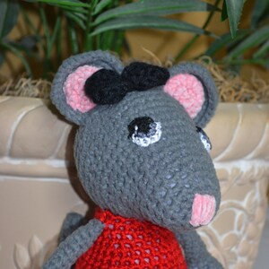 May include: A gray crocheted mouse wearing a red crocheted dress with a black bow. The mouse has pink ears and a white eye with a black pupil.