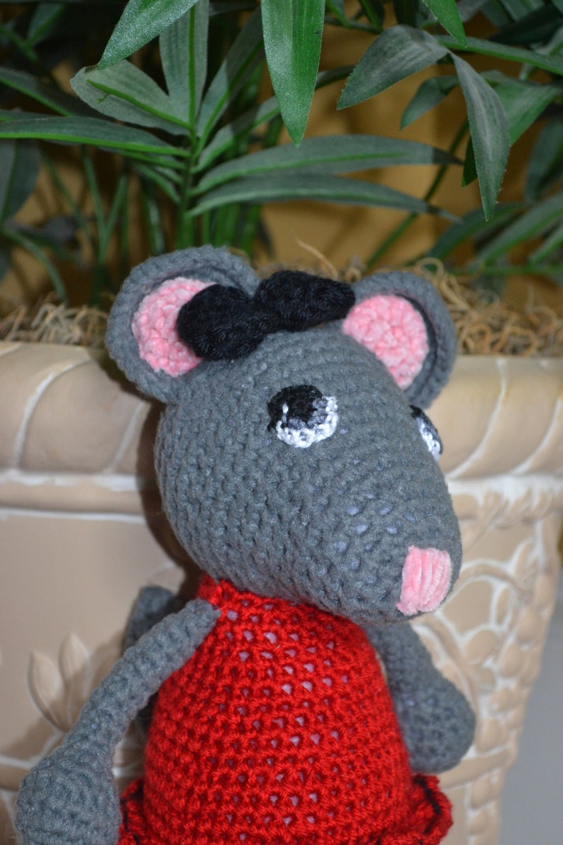 May include: A gray crocheted mouse wearing a red crocheted dress with a black bow. The mouse has pink ears and a white eye with a black pupil.