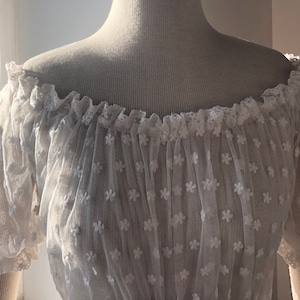 May include: White off-the-shoulder blouse with a delicate floral pattern and lace trim. The blouse has a loose, flowing fit and is made of a sheer fabric.