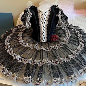 May include: A black and gold ballet tutu with a velvet bodice and lace detailing. The bodice has a white panel with criss-cross black lacing. The tutu has multiple layers of tulle with gold and red accents, and a red rose.