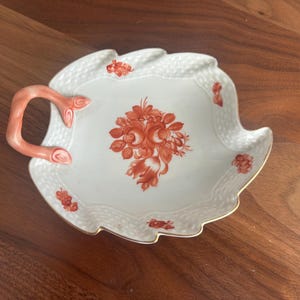 May include: White leaf-shaped serving dish with a coral handle and floral design. The dish features a detailed, textured rim and a gold-colored edge. The central floral arrangement and smaller accents are in a coral hue.