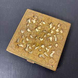 May include: A vintage gold-toned compact with a floral filigree design. The square compact features an intricate pattern of flowers and leaves, with a lighter-colored interior visible through the cut-out design. The metal has a slightly aged appearance.