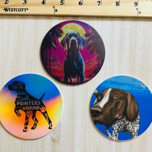 Puede incluir: Tres posavasos redondos con diferentes diseños de Braco Alemán de Pelo Corto. Un posavasos tiene una silueta en blanco y negro de un braco con el texto "La vida es mejor con Bracos alrededor". Otro posavasos tiene una ilustración colorida de un braco frente a una puesta de sol con palmeras. El tercer posavasos tiene una ilustración realista de un braco frente a un fondo azul con burbujas.