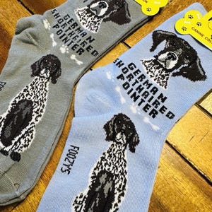 Può includere: Due paia di calzini, uno grigio e uno azzurro chiaro, ciascuno con un disegno di Bracco Tedesco. I calzini hanno la scritta "GERMAN SHORTHAIRED POINTER" e il marchio "FOOZYS" è visibile.