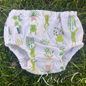 May include: White baby bloomers with a green frog and potted plant print. The print features frogs in various poses and potted plants with green leaves.